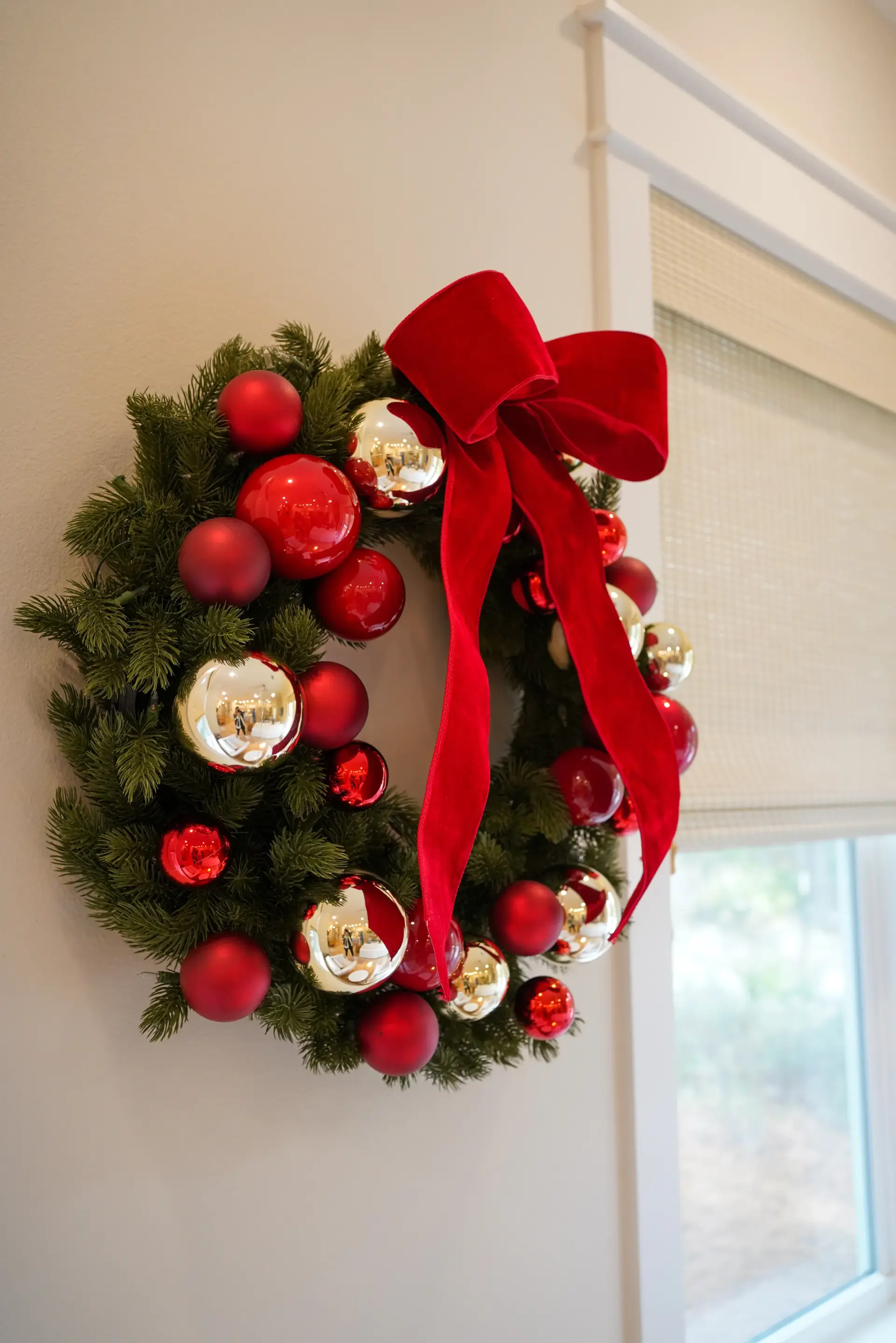 A Christmas wreath with red ornaments and a red bow beside a window